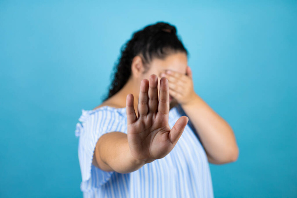 Jonge mooie vrouw met krullend haar over geïsoleerde blauwe achtergrond bedekken ogen met handen en doen stop gebaar met droevige en angst uitdrukking. Beschaamd en negatief concept. - Foto, afbeelding
