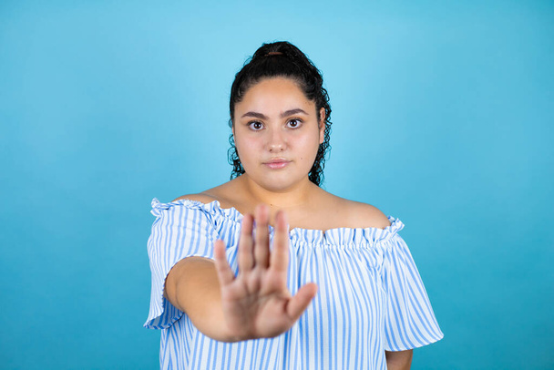Young beautiful woman with curly hair over isolated blue background serious and doing stop sing with palm of the hand. - Photo, Image