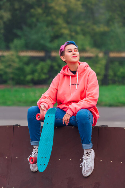 Young girl with short multicolored hair and nose piercing dressed in pink hoodie sits with skateboard in skatepark during morning fog. Portrait of a tomboy with plastic skateboard - 写真・画像