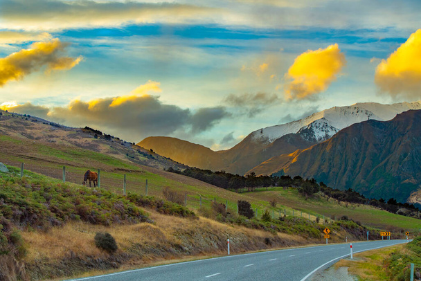 Vista sulle montagne e sulla strada a Wanaka Otago, Newzealand quando il sole tramonta. C'era una bella nuvola gialla e un cavallo stava mangiando erba sulla collina. - Foto, immagini