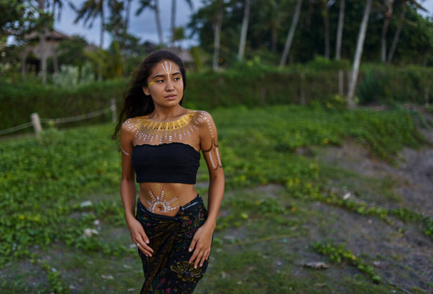 A young, beautiful, ethnic girl with drawings on her body against the background of palm trees and green grass. - Photo, Image