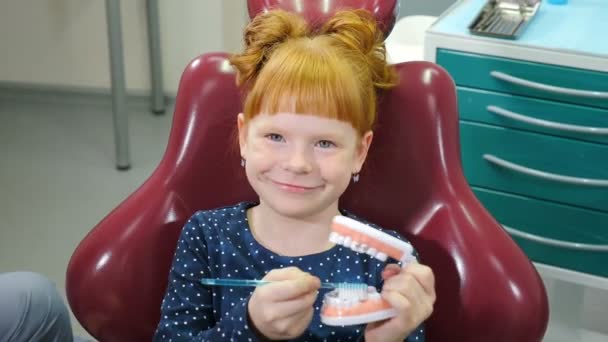 Pediatrik dişçilik konsepti. Dişçi koltuğundaki sevimli küçük kız. Güzel kızıl saçlı kız diş hekimliğinde eğleniyor, diş modelleriyle oynuyor fırçayla diş temizliyor. 4k video - Video, Çekim
