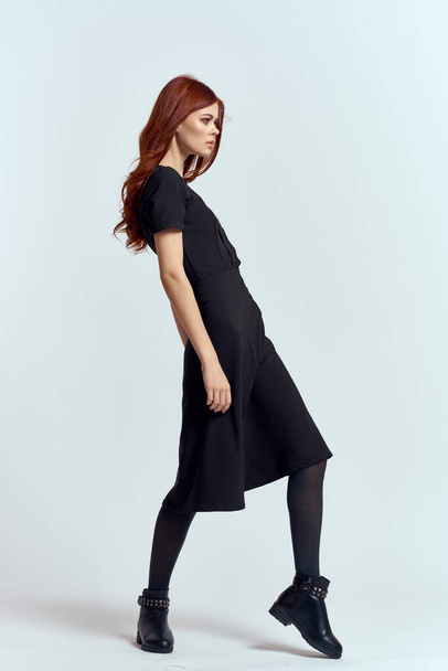 A woman in a black dress on a light background and pantyhose shoes red hair and pose in full growth - Fotografie, Obrázek