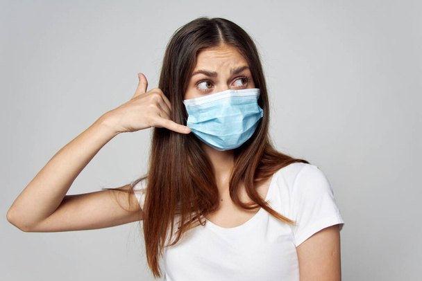 Chica con una máscara médica sostiene dos dedos cerca de la cara en una camiseta blanca  - Foto, imagen