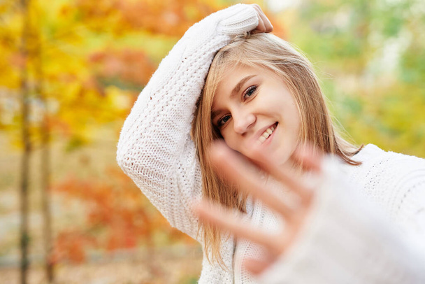 Vonzó fiatal mosolygós szőke modell lány fehér pulóver pózol háttér őszi park. Teenager portré, modell megjelenés vagy őszi őszi koncepció - Fotó, kép