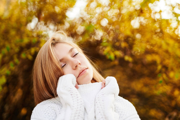 Ονειρεμένη ξανθιά μοντέλο κορίτσι σε λευκό πουλόβερ ποζάρουν με φόντο το φθινόπωρο πάρκο. Κοντινό πορτραίτο όμορφου κοριτσιού με κλειστά μάτια. Teenager πορτρέτο, μοντέλο εμφάνιση ή φθινόπωρο έννοια πτώση - Φωτογραφία, εικόνα