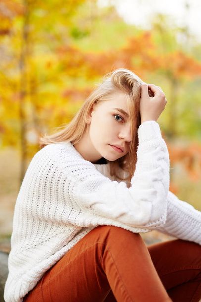 Menina modelo adolescente loira bonita em camisola branca e jeans laranja sentado com fundo do parque de outono e olhando diretamente para a câmera. Teen retrato, modelo olhar livro ou outono outono outono conceito - Foto, Imagem
