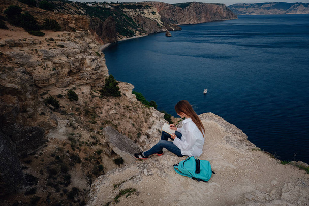 魅力的な女性の海の景色と山の端には、本を読んで座っている。オープンエアでの自己教育。日記にメモを書き, - 写真・画像