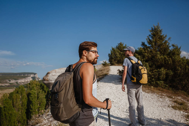 Twee mannen lopen op rotsachtige helling dragen rugzakken met behulp van trekking Sticks. Bergen vallei Uitzicht naast de mensen. Zomer reizen naar de natuur. Wandelaar op de achtergrond. - Foto, afbeelding