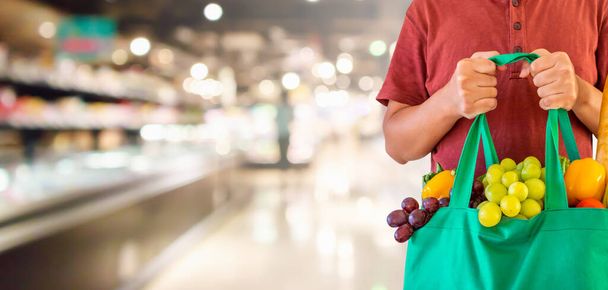 Müşteri, süpermarket koridorunun üzerinde meyve ve sebzelerle yeniden kullanılabilir yeşil alışveriş torbası bulundurur. Odaklanmış ürün rafları, Bokeh light arka plan. - Fotoğraf, Görsel