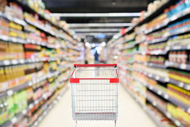 Empty shopping cart with abstract blur supermarket discount store aisle and sauce seasoning product shelves interior defocused background - Фото, изображение
