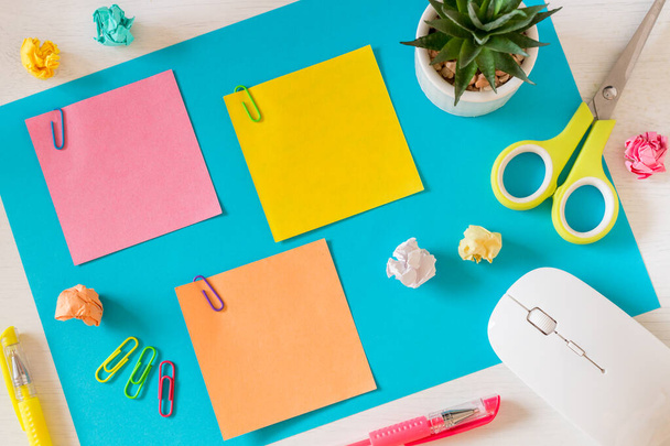 A large, blue card on the desk and three small colored sheets with space for text. Office and school accessories, computer mouse, pens, crumpled paper balls, scissors, mini flower in a pot, staples.  - Photo, Image