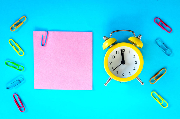 Blue background, pink card with an office staple and space for an inscription. Next to it, an alarm clock is set for an hour, five to midnight. Concept for business and office purposes - 写真・画像