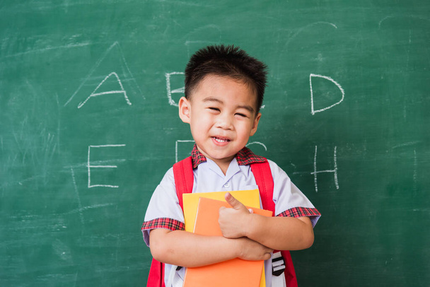 Back to School. Happy Asian funny cute little child boy kindergarten in student uniform with school bag and books smile show finger thumb up on green school blackboard, First time to school education - 写真・画像