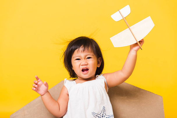 Happy Asian beautiful funny baby little girl smile wear pilot hat play with toy cardboard airplane wings fly hold plane toy, studio shot isolated yellow background, Startup freedom concept - Фото, зображення