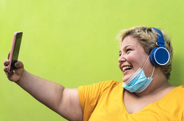 Plus size woman taking selfie with mobile smartphone and listening to music with wireless headphones while wearing face protective mask to avoid corona virus infection - Health and technology concept - Foto, Imagem