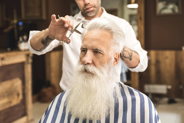 Hairdresser making stylish haircut for a handsome old man in the barbershop - Foto, Imagen