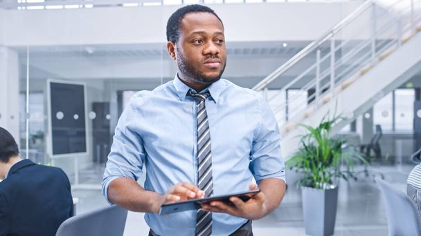 Handsome Young Black Manager in a Shirt Walking Pass His Business Colleagues with a Tablet. Diverse and Motivated Business People Work on Computers in Modern Open Office. - Photo, Image