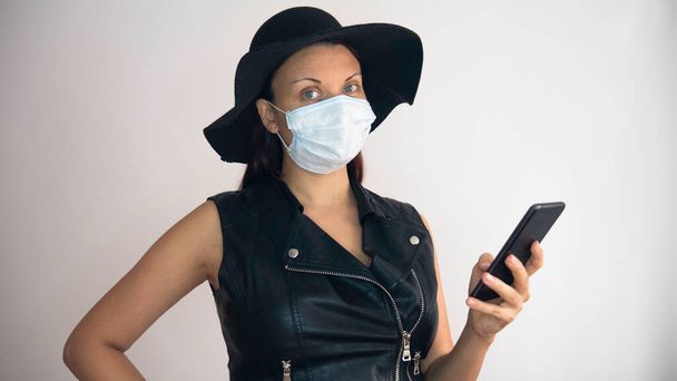 黒い帽子をかぶった黒い革のベストの女性と彼女の顔に医療マスクは白い背景に携帯電話を使用しています. - 写真・画像