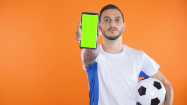 Fußballfan in weißblauem Hemd und Ball zeigen grünen Bildschirm Smartphone orange Hintergrund  - Filmmaterial, Video