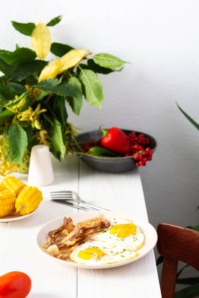 Сытный завтрак на столе, жареные яйца с беконом и вареной кукурузой и овощами, осенние листья в вазе - Фото, изображение
