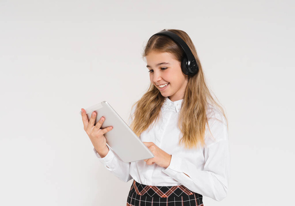 Bonito sorridente menina adolescente em uniforme escolar com fones de ouvido e tablet em suas mãos isolado no fundo branco - Foto, Imagem
