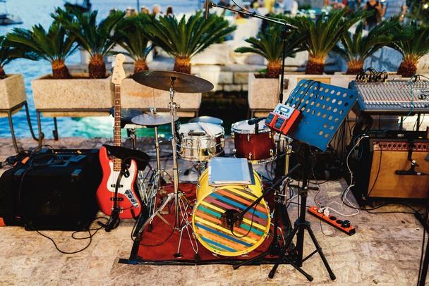 Μια πλατφόρμα για μουσικούς με μουσικά όργανα, drum kit, κιθάρα και κονσόλα ηχητικών μηχανικών στο φόντο ενός γιορτινού τραπεζιού δίπλα στο νερό. - Φωτογραφία, εικόνα