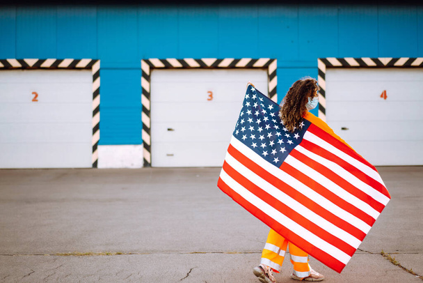 African American woman outdoors, wearing a protective mask against coronavirus and holding a USA flag. - Foto, Bild