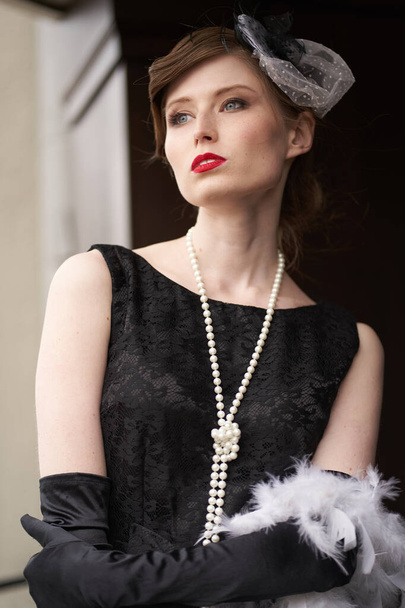 Όμορφη μελαχρινή γυναίκα με καπέλο, σε μαύρο ρετρό φόρεμα με κόκκινα χείλη. Στέκεται και κοιτάζει κατευθείαν στην κάμερα, στο βάθος μιας ξύλινης πόρτας                                - Φωτογραφία, εικόνα