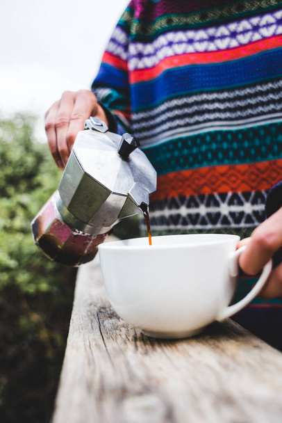 Eine Frau gießt Kaffee in eine weiße Tasse aus einer Geysir-Kaffeemaschine, Morgens Heißgetränk in der Natur, Mädchen kocht Kaffee auf der Veranda eines Holzhauses. - Foto, Bild