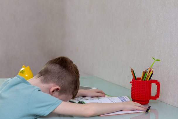 Κουρασμένο θλιμμένο αγόρι που σπουδάζει στο σπίτι του στην καραντίνα. Ένα θλιμμένο αγόρι που διαβάζει στο διαδίκτυο. Αποκοιμηθείτε στο γραφείο ενώ κάνετε τα μαθήματά σας. - Φωτογραφία, εικόνα