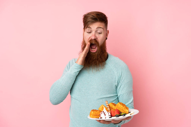 Redhead man with long beard holding waffles over isolated pink background with surprise and shocked facial expression - Foto, Bild