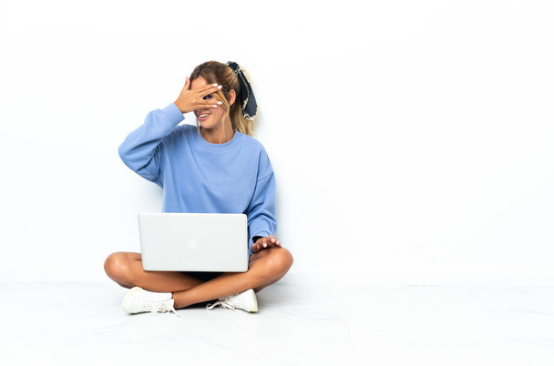 Młoda blondynka urugwajska dziewczyna z laptopem odizolowana na białym tle, okrywająca oczy dłońmi i uśmiechnięta - Zdjęcie, obraz