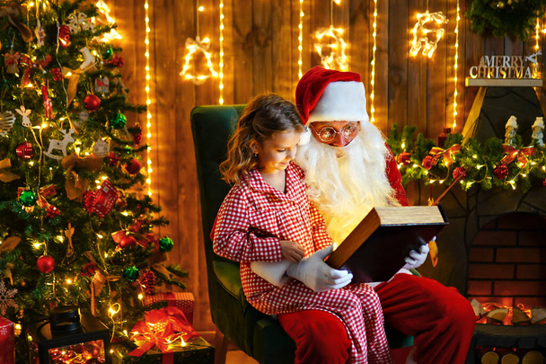 Santa Claus open and read magic book with little cute amazed girl in pajama - Fotó, kép