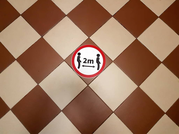 Un letrero de piso redondo con símbolos de dos personas y letras que dicen 2m dice 'por favor, asegure el distanciamiento social' en un piso de baldosas - Foto, imagen