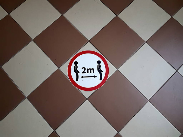 Круглый вывеска с символами двух человек и надписью 2m говорит: "Пожалуйста, убедитесь, что социальное дистанцирование" на плитке пола - Фото, изображение
