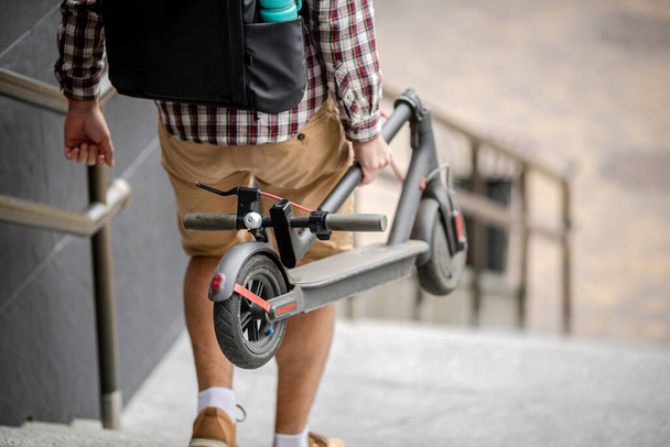 Jonge blanke man in casual kleding met een rugzak draagt een opvouwbare elektrische scooter in zijn hand de trap op van een kantoorgebouw. Man draagt E-Scooter in gevouwen positie van het werk. - Foto, afbeelding