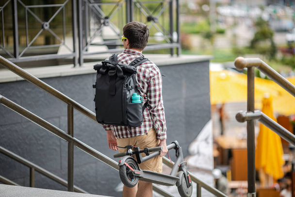 Katlanmış bir E-scooter taşıyan bir adam binanın yanındaki merdivenlerde. Çevresel şehir taşımacılığı, modern şehir konsepti. Ekolojik teknolojik yaşam tarzı. Katlanmış Konumda Elektrikli Scooter Taşıyıcı. - Fotoğraf, Görsel