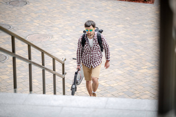 男は近代的な建物の近くの都市の階段を電動スクーターの上に折り畳まれた歩く。生態系の技術ライフスタイル。Eモビリティ。電動スクーターを運ぶ者は、職を畳んだ。エコ都市交通. - 写真・画像
