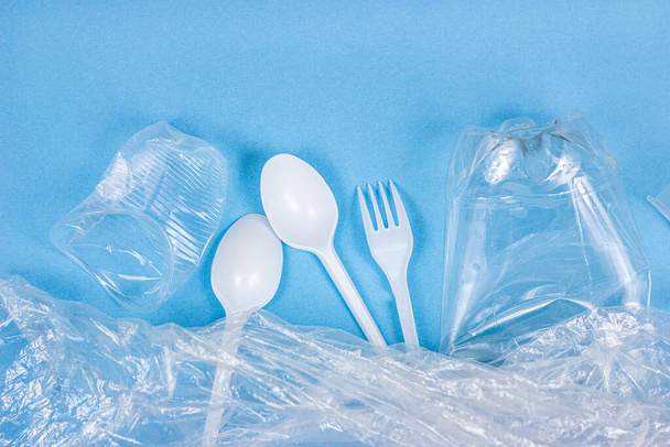 Top view of crushed plastic spoons, forks and cups as a disposable waste with copy space on bright blue background. Environmental pollution and litter recycling concept. - Photo, Image