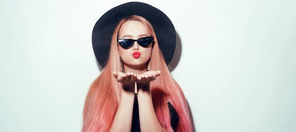 Девушка с розовыми волосами в очках и шляпой в черной одежде. Черная пятница. Модель шлет поцелуй - Фото, изображение