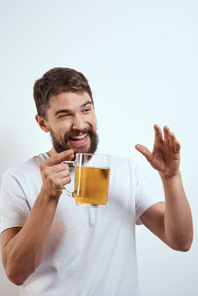 Hafif arka planda bira fincanı olan sakallı bir adam. Beyaz tişörtlü, alkollü bir içkinin görüntüsü. - Fotoğraf, Görsel