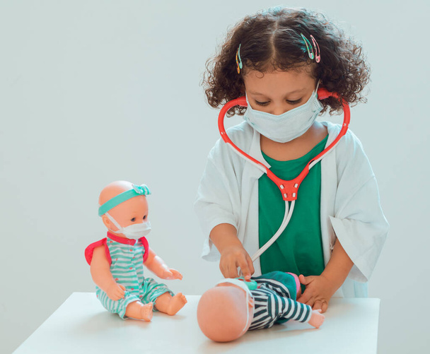 A little girl playing the doctor listens to the heart of her patient with a stethoscope while another patient watches, all wear protective masks - Zdjęcie, obraz