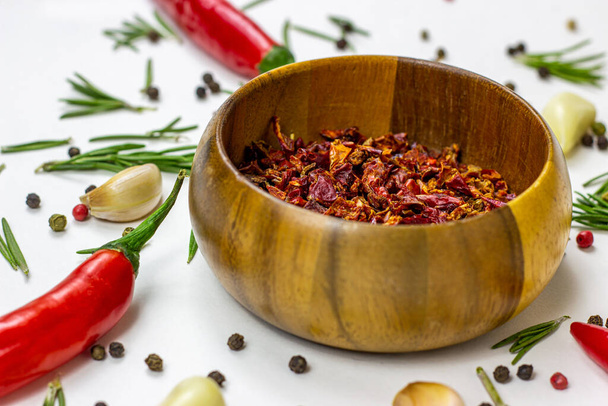 Dry red chili pepper slices in a round wooden bowl with peppercorns, garlic and rosemary on white food background. - Photo, Image