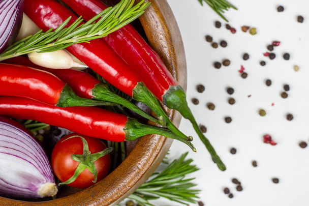 Widok na góry warzyw letnich: cebula, czosnek, pomidor, czerwony pieprz chili, rozmaryn w brązowej drewnianej misce i papryka na białym tle stołu. - Zdjęcie, obraz