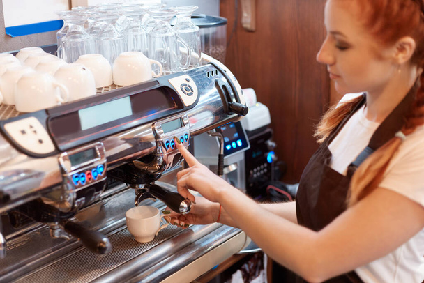 Professional barista preparing coffee using coffee maker, young woman making coffee, red haired female working in coffee shop, wearing white casual t shirt and brown apron. - Fotoğraf, Görsel