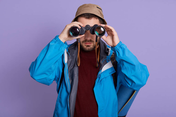 Man Hiking and birdwatching, looking through binoculars, wearing blue jacket, hat and warm sweater, posing isolated over lilac background, tourist hiking. - Valokuva, kuva