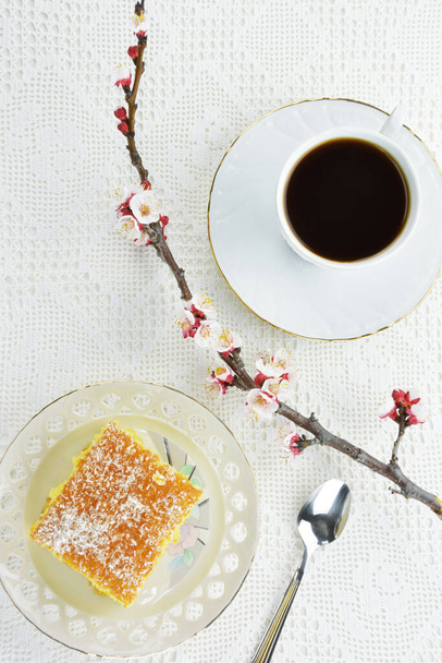 Ravani con coco en copos en la parte superior y empapado en almíbar, una taza de café negro y una rama con flores en el fondo de la estera de encaje - Foto, Imagen