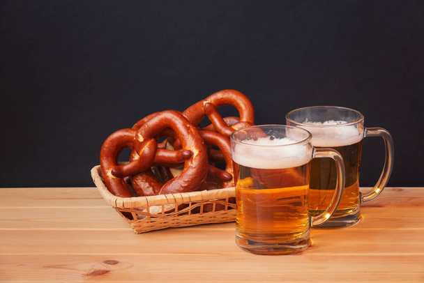 Dos tazas de cerveza y cesta de mimbre con pretzels suaves sobre mesa de madera sobre fondo oscuro. Enfoque selectivo. Ver con espacio de copia. - Foto, imagen