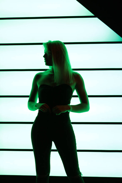 Club style photo of girl in a black corset and jeans.  Set is a lit with a green color. Picture has dark  tone. Wall of neon has green color.  - Foto, afbeelding